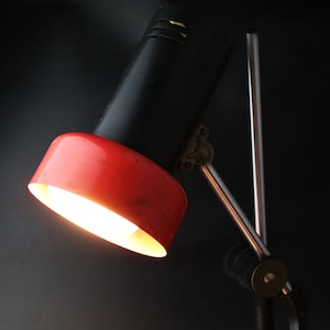 May include: A black and red desk lamp with a metal arm and a red shade. The lamp is turned on and the light is shining.