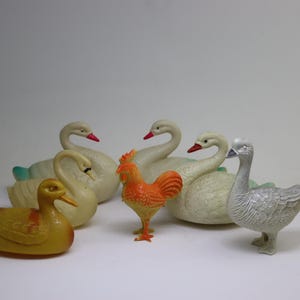 May include: A collection of five vintage plastic toy farm animals, including a rooster, a duck, and three swans. The rooster is orange and white, the duck is yellow, and the swans are white.