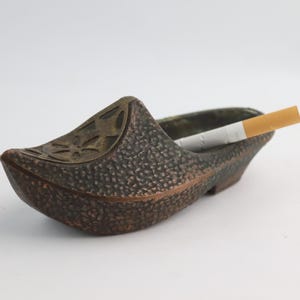 May include: A bronze-coloured ashtray in the shape of a Dutch clog, featuring a textured finish and a floral design on the front. A lit cigarette is placed in the ashtray, set against a white backdrop.