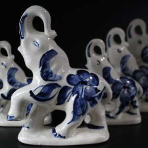 May include: A collection of white ceramic elephant figurines with blue floral and leaf designs. The elephants are standing on a base and have their trunks raised. The figurines are arranged in a row, with some in focus and others blurred.