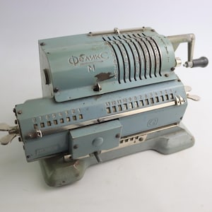 Puede incluir: Calculadora mecánica azul-gris vintage con múltiples diales y palancas numéricas. La calculadora tiene una manivela y una serie de teclas numéricas. El texto "ФЕЛИКС M" es visible en la parte superior.