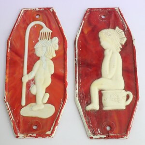 May include: Two vintage bathroom signs with a red background and white figures. The sign on the left features a figure holding a shower head, while the sign on the right features a figure sitting on a toilet.