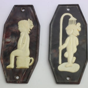 May include: Two vintage bathroom signs with a black and white marbled background. The signs feature a white raised image of a child. One sign shows a child sitting on a toilet and the other shows a child holding a shower head.