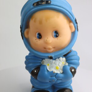 K&ouml;nnte beinhalten: Ein blauer Plastik-Astronautenspielzeug, das einen Strau&szlig; wei&szlig;er Blumen h&auml;lt. Der Astronaut hat ein rundes, l&auml;chelndes Gesicht mit blauen Augen und Sommersprossen.