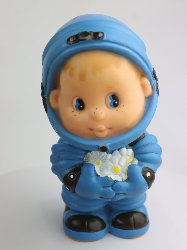 K&ouml;nnte beinhalten: Ein blauer Plastik-Astronautenspielzeug, das einen Strau&szlig; wei&szlig;er Blumen h&auml;lt. Der Astronaut hat ein rundes, l&auml;chelndes Gesicht mit blauen Augen und Sommersprossen.