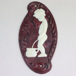 May include: Oval-shaped, burgundy-colored wall plaque with a white relief of a boy urinating into a small pot. The plaque has a decorative border and two holes at the top for hanging.