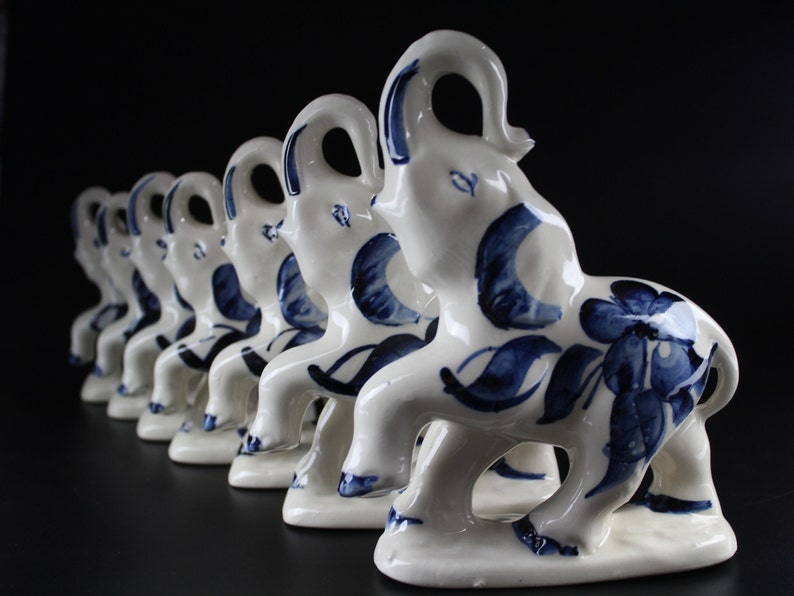 May include: A row of white ceramic elephant figurines with blue floral and leaf designs. The elephants are in a line, each with a raised trunk and a decorative pattern on their bodies. The background is black.