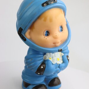 K&ouml;nnte beinhalten: Ein blaues Plastik-Astronautenspielzeug, das einen Strau&szlig; wei&szlig;er Blumen h&auml;lt. Das Spielzeug hat einen blauen Helm, einen blauen Raumanzug und schwarze Stiefel. Das Spielzeug hat ein freundliches Gesicht mit blauen Augen und Sommersprossen.
