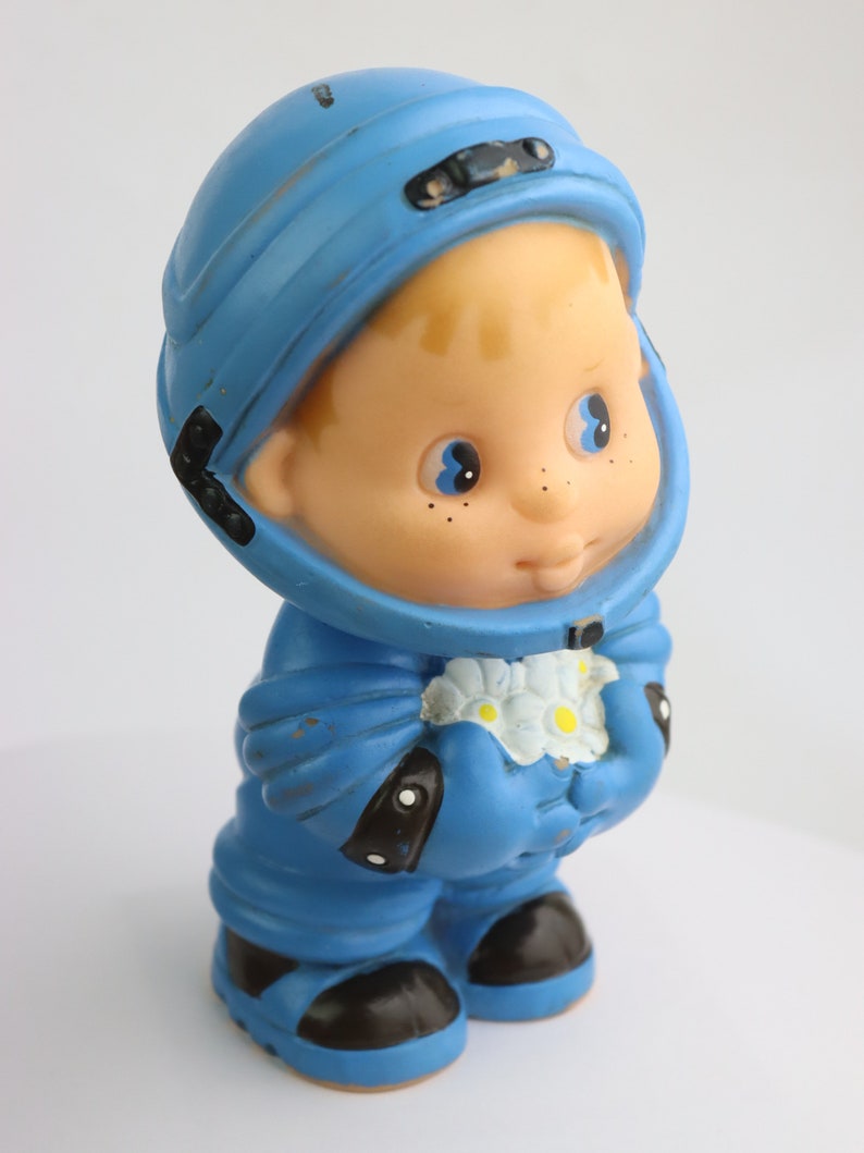 K&ouml;nnte beinhalten: Ein blaues Plastik-Astronautenspielzeug, das einen Strau&szlig; wei&szlig;er Blumen h&auml;lt. Das Spielzeug hat einen blauen Helm, einen blauen Raumanzug und schwarze Stiefel. Das Spielzeug hat ein freundliches Gesicht mit blauen Augen und Sommersprossen.