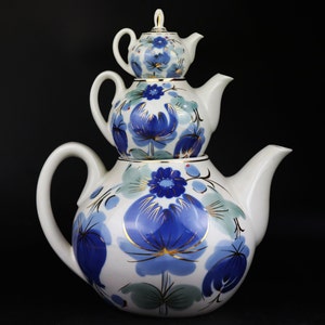 Könnte beinhalten: Ein Set aus drei weißen Porzellan-Teekannen mit blauen und goldenen Blumendesigns. Die Teekannen sind übereinander gestapelt, wobei die kleinste Teekanne oben liegt.
