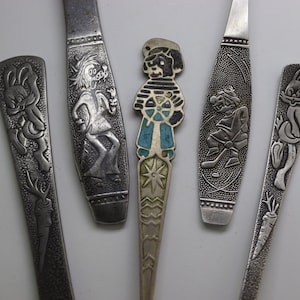 May include: A selection of vintage silver-coloured spoons with embossed cartoon character designs. The spoons feature characters like Bugs Bunny, a wolf-like figure, a sailor, and a hockey-playing character. The central spoon has a sailor design with blue and turquoise accents.