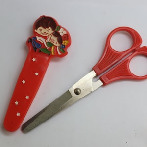 May include: Red plastic scissors with a cartoon image of two children on the handle. The scissors have a silver blade.