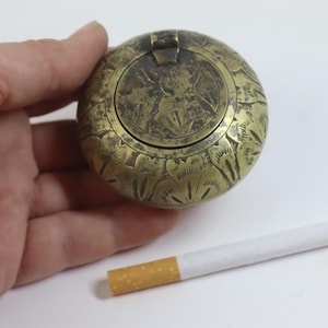 May include: A round, metallic brass-toned container featuring an engraved floral pattern on the exterior and a hinged lid. The object is shown next to a standard cigarette for scale, highlighting its compact, circular design and weathered finish.