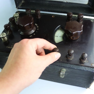 May include: A vintage black electrical testing device with brown knobs and a gauge. The device is housed in a black case with a hinged lid. A hand is turning one of the knobs.