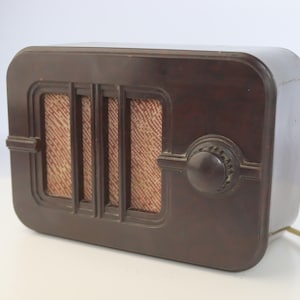 Puede incluir: Una radio marrón vintage con forma rectangular y bordes redondeados. La parte frontal presenta una rejilla de altavoz con barras verticales y una tela texturizada. Una perilla circular está en el lado derecho, y un cable de alimentación sale de la base.