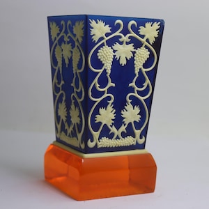 May include: A decorative blue vase with a cream-coloured grape vine design. The vase sits on an orange base. The vase is a square shape and is made of a glossy material.