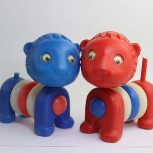 Könnte beinhalten: Zwei Vintage-Spielzeugtiere, eines blau und eines rot, jeweils mit einem segmentierten Körper aus blauen, weißen und roten Ringen. Die Spielzeuge haben runde Köpfe mit großen Augen und kleinen Ohren, wahrscheinlich aus der Mitte des 20. Jahrhunderts.