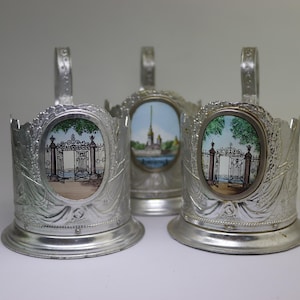May include: Three silver-colored cup holders with ornate designs. Each holder features a handle and an oval-shaped, colorful enamel insert depicting a landscape scene. The cup holders are likely vintage or antique.
