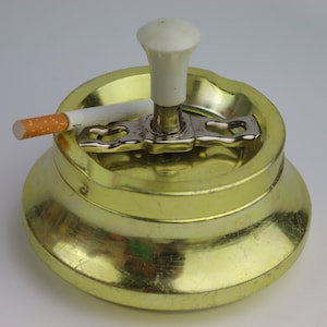 Puede incluir: Un cenicero de metal amarillo con una tapa de plástico blanco y un porta cigarrillos de metal. Un cigarrillo descansa sobre el cenicero.