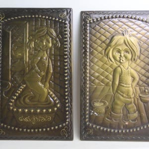 May include: Two vintage metal wall plaques. One depicts a child in a bathtub, the other a child standing with a bucket. Both are embossed with a gold-toned finish and framed in a dark brown border with a beaded edge.