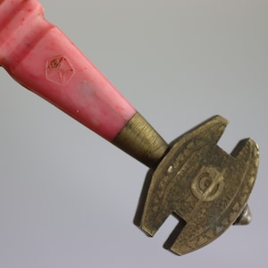 May include: A pink plastic handle attached to a brass glass cutter with a circular blade and a small, pointed tip. The cutter has a decorative design with a letter "G" in the centre.