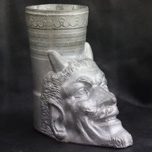 May include: A silver-colored, devil-themed drinking mug. The mug features a detailed devil face on the side, with horns, a pointed beard, and a sinister grin. The top of the mug has a decorative band. The mug is made of a metallic material.