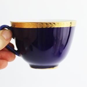 May include: A cobalt blue teacup with a gold rim and a white interior. The cup is held in a hand.