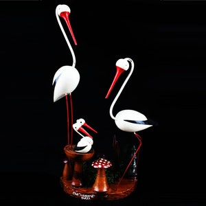 May include: A wooden sculpture of a family of storks. The storks are white with red beaks and black markings. The sculpture is set on a wooden base with a mushroom and a small tree.