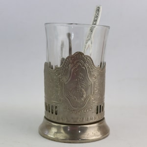 Puede incluir: Calientavasos de metal plateado con un diseño ornamentado, que sostiene una taza de té de vidrio transparente. Un billete de dólar está metido en la taza.