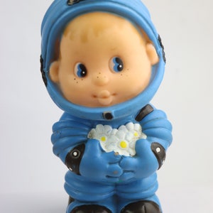 K&ouml;nnte beinhalten: Ein blaues Plastik-Astronautenspielzeug, das einen Strau&szlig; wei&szlig;er Blumen h&auml;lt. Der Astronaut hat ein rundes, l&auml;chelndes Gesicht und einen blauen Helm.