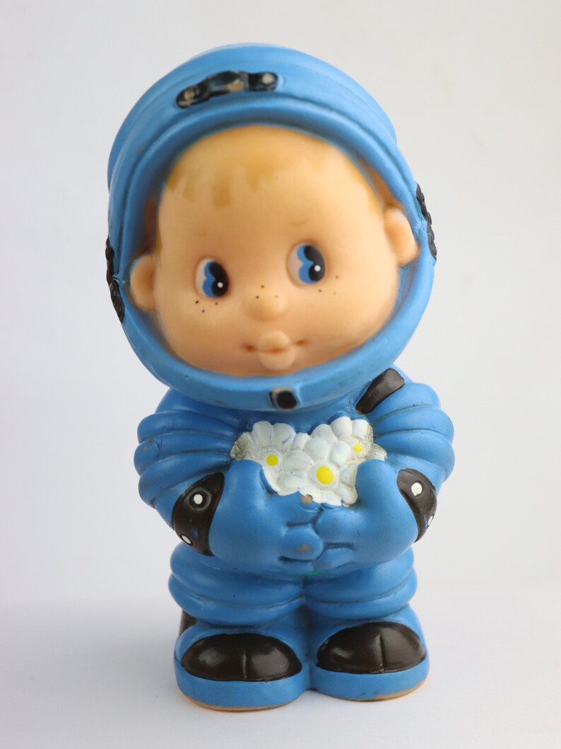 K&ouml;nnte beinhalten: Ein blaues Plastik-Astronautenspielzeug, das einen Strau&szlig; wei&szlig;er Blumen h&auml;lt. Der Astronaut hat ein rundes, l&auml;chelndes Gesicht und einen blauen Helm.