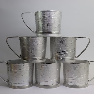 Könnte beinhalten: Sechs silberfarbene Metallbecher gestapelt. Jeder Becher hat einen Henkel und ist mit einem Himmelsdesign aus Sternen, Planeten und geschwungenen Linien verziert. Die Becher haben eine strukturierte Oberfläche und einen Sockel.