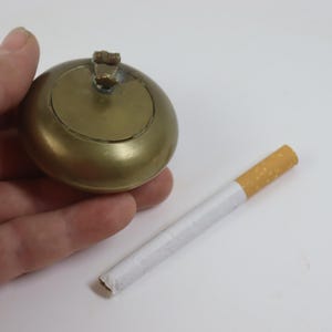 May include: A small, round, brass-colored metal ashtray with a hinged lid mechanism, positioned next to a standard white cigarette with a tan filter. The ashtray has a smooth, polished surface and a circular central opening for ash disposal.