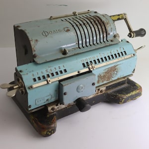 Puede incluir: Calculadora mecánica vintage azul y gris con manivela y diales numéricos. La calculadora muestra signos de desgaste, con óxido y desconchones de pintura. El texto "Фоликс" es visible en la parte superior.