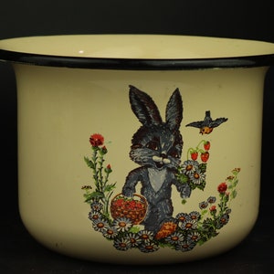 May include: A white enamelware mug with a black rim features a cartoon bunny holding a basket of strawberries. The bunny is surrounded by flowers and a small bird.