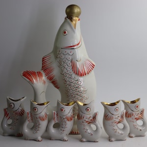 Puede incluir: Una jarra de cerámica blanca con forma de pez con un tapón dorado y seis vasos de chupito a juego. El pez tiene detalles rojos y dorados. Los vasos de chupito tienen forma de pez con bordes dorados.