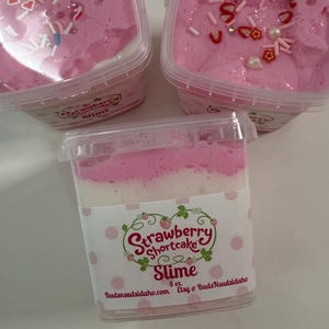 May include: Three square clear plastic containers filled with pink and white layered slime. The tops are decorated with small strawberry, heart, and pearl-shaped charms. Each container features a label reading Strawberry Shortcake Slime 8 oz.