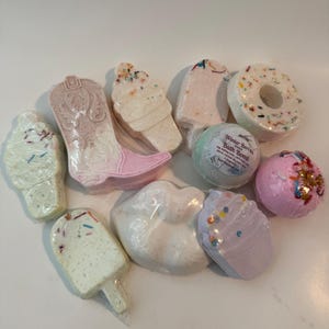 May include: A collection of colorful bath bombs in various shapes, including an ice cream cone, a cowboy boot, a donut, and a pair of lips. The bath bombs are decorated with sprinkles and other details.