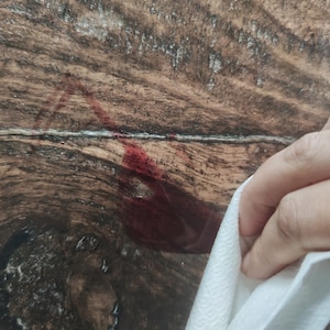 May include: A close-up of a dark wood surface with a red stain. A white paper towel is being used to wipe the stain.