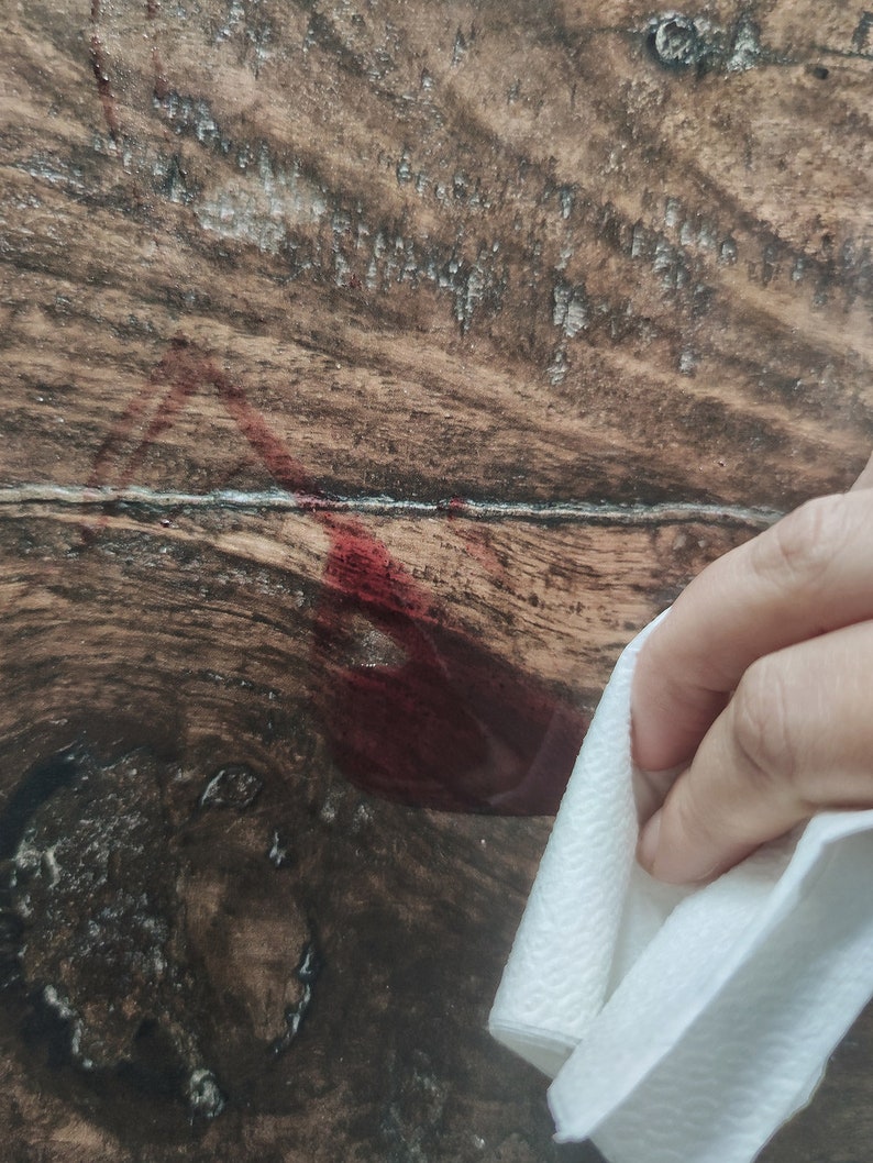 May include: A close-up of a dark wood surface with a red stain. A white paper towel is being used to wipe the stain.