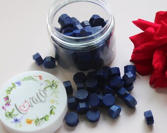 May include: A clear jar filled with navy blue wax seal beads, with some spilled out. A white lid with a floral wreath and the word "love" is next to the jar. A red rose is in the background. Ideal for crafting and stationery.