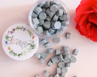 May include: A clear plastic container filled with light blue wax seal beads. The lid, decorated with a floral wreath and the word "love", is next to the container. A red rose is in the background. The beads are scattered around the container.