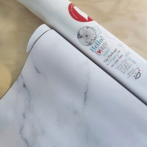 May include: A white tube with a red heart and the text "Hello! Walo" on it. The tube is rolled up and the text "This Package is Happy to see you too." is visible. The tube is on a white marble surface.