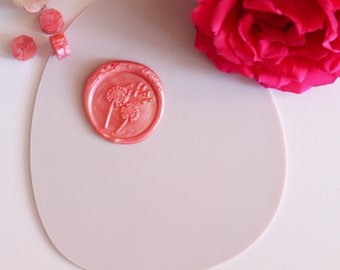 May include: A pink wax seal with a dandelion design, surrounded by loose wax pieces and a gold stamp. A vibrant red rose is in the background. The seal is on a light pink oval-shaped surface.