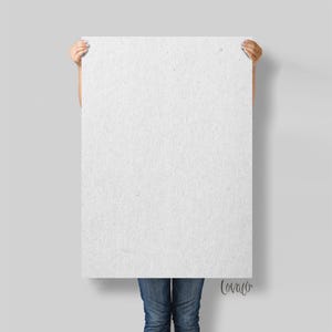 Könnte beinhalten: Ein großes, rechteckiges weißes Papierblatt mit strukturierter Oberfläche wird von einer Person gehalten. Die Person trägt eine blaue Jeans. Das Papier ist leer, bereit für Text oder Kunst. Der Hintergrund ist hellgrau.
