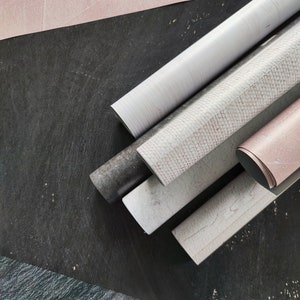 May include: A close-up of five rolls of decorative contact paper. The rolls are arranged in a fan shape on a black surface. The contact paper has a variety of patterns, including a white woodgrain, a white and pink speckled pattern, and a solid pink.