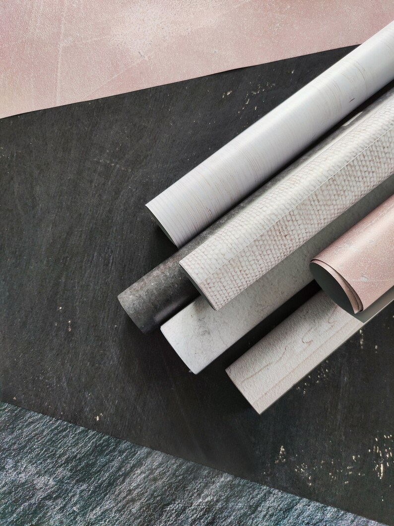 May include: A close-up of five rolls of decorative contact paper. The rolls are arranged in a fan shape on a black surface. The contact paper has a variety of patterns, including a white woodgrain, a white and pink speckled pattern, and a solid pink.