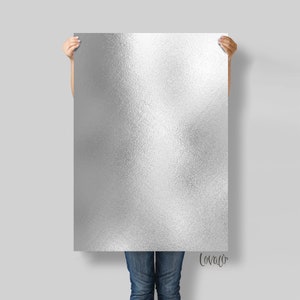 May include: A silver metallic poster with a textured, brushed finish. The poster is being held by a person, but only their hands and jeans are visible.