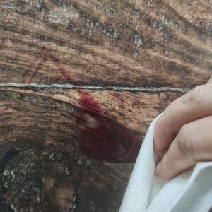 May include: A hand holding a white paper towel is wiping up a red liquid spill on a wooden surface. The wood has a natural grain and a dark brown color.