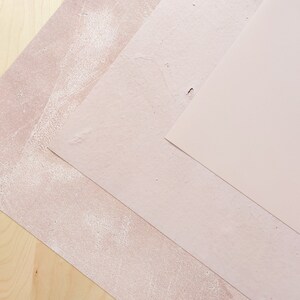 Pu&ograve; includere: Tre pezzi di carta testurizzata in tonalit&agrave; rosa, disposti in diagonale su una superficie di legno chiaro.